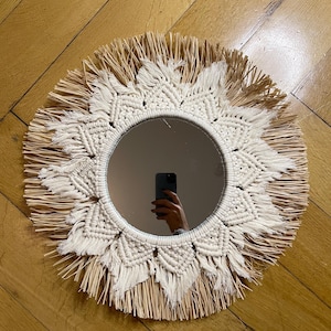 Könnte beinhalten: Ein runder Spiegel mit einem gewebten Makramee-Rahmen und einem natürlichen Strohrand. Der Spiegel ist von einem weißen Makramee-Muster umgeben.