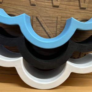 May include: Three wooden shelf brackets in a wave shape, stacked on top of each other. The top bracket is light blue, the middle bracket is black, and the bottom bracket is white.
