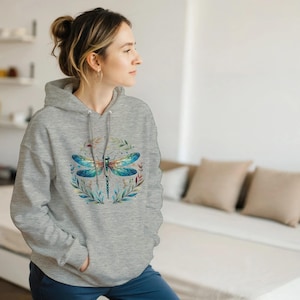 Può includere: Una felpa con cappuccio grigio chiaro con un design di libellula ad acquerello. La libellula ha ali blu e turchese, con un corpo dettagliato, circondato da una ghirlanda di foglie e fiori. La felpa ha un cappuccio con coulisse e una tasca frontale.