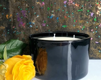 May include: A glossy black candle in a round container with two wicks. A yellow rose and green leaf are in the foreground. The background is a dark canvas with colorful paint splatters. The candle has a white wax.