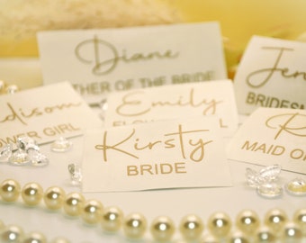 May include: White cards with gold lettering for a wedding party. The cards read "Kirsty Bride", "Emily Flower Girl", "Madison Flower Girl", "Diane Mother of the Bride", "Jenna Bridesmaid", and "Rachel Maid of Honor".