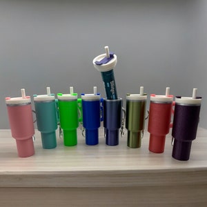 Puede incluir: Una fila de diez tazas de viaje coloridas con una tapa blanca y una pajita. Las tazas son de varios colores, incluyendo rosa, azul, verde y morado. Las tazas tienen una banda plateada alrededor del medio. Un tubo de plástico transparente con una tapa blanca y una pajita está de pie en el centro de la imagen.