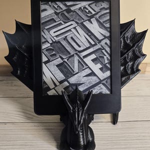 Könnte beinhalten: Ein schwarzer E-Reader wird von einem 3D-gedruckten Drachenständer gehalten. Der Drache hält den E-Reader in seinen Vorderpfoten. Der Bildschirm des E-Readers zeigt ein schwarz-weißes Muster aus Buchstaben.