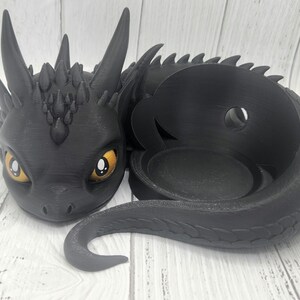 Könnte beinhalten: Schwarzer drachenförmiger Kerzenhalter mit einer runden, offenen Oberseite für eine Kerze. Der Drache hat gelbe Augen und einen strukturierten, schuppigen Körper.