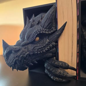 Könnte beinhalten: Schwarzer Drachenkopf-Buchstütze mit strukturiertem, schuppigem Design. Das Auge des Drachen ist leuchtend gelb. Die Buchstütze hält ein Buch mit seiner Klaue.