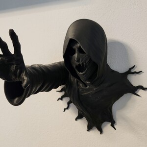 Könnte beinhalten: Eine schwarze, 3D-gedruckte Wandskulptur einer Kapuzenfigur mit einem bedrohlichen Ausdruck. Die Hand der Figur ist ausgestreckt, als würde sie aus der Wand greifen.