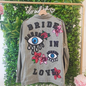 Peut inclure: Une veste utilitaire verte avec les mots "Bride in Love" en sequins noirs. La veste a un grand œil bleu avec des paillettes et des fleurs roses au dos.