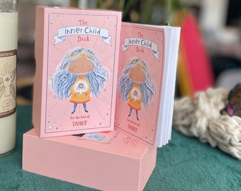 May include: A pink box with the text "The Inner Child Deck" and a cartoon illustration of a girl with blue hair. The box is stacked on top of another pink box with the text "for the love of TAROT".