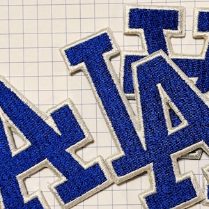 May include: Embroidered patches displaying the letters "A" and "T" in blue with a white border. The patches have a textured finish and are arranged on a grid-lined surface. These patches are suitable for various applications.