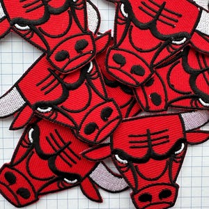 Könnte beinhalten: Haufen roter, gestickter Bullenkopf-Patches mit schwarzen und weißen Details. Die Patches zeigen einen Bullenkopf mit Hörnern, Augen und Gesichtszügen. Die Patches sind wahrscheinlich für Kleidung oder Accessoires gedacht.