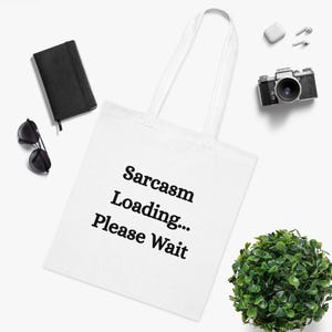 May include: White tote bag with the text "Sarcasm Loading... Please Wait" in black. The bag is surrounded by a black notebook, sunglasses, a camera, earbuds, and a small green plant.