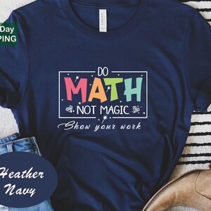May include: Heather navy t-shirt with the text "DO MATH NOT MAGIC Show your work" in a colorful design. The shirt has a crew neck and short sleeves. The shirt is made of a soft material.