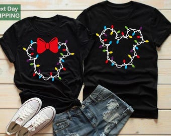 Op de afbeelding: Twee zwarte T-shirts met een Mickey Mouse-hoofdontwerp gemaakt van kerstverlichting. Eén shirt heeft een rode strik. Witte sneakers en een spijkerbroekshort staan op de voorgrond. Voor koppels of bijpassende outfits.