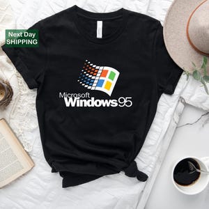 Puede incluir: Camiseta negra con el logotipo de Microsoft Windows 95 en blanco, rojo, azul y amarillo. La camiseta tiene cuello redondo y mangas cortas. El texto "Microsoft Windows 95" est&aacute; impreso debajo del logotipo. La camiseta se muestra sobre una superficie blanca.
