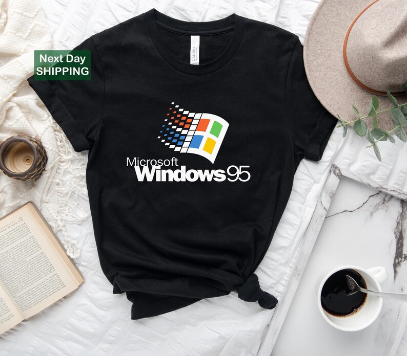Puede incluir: Camiseta negra con el logotipo de Microsoft Windows 95 en blanco, rojo, azul y amarillo. La camiseta tiene cuello redondo y mangas cortas. El texto "Microsoft Windows 95" est&aacute; impreso debajo del logotipo. La camiseta se muestra sobre una superficie blanca.