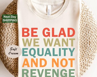 Puede incluir: Una camiseta de cuello redondo color crema con el texto BE GLAD WE WANT EQUALITY AND NOT REVENGE impreso en líneas apiladas de letras mayúsculas en rojo, verde oliva, azul verdoso y naranja. La camiseta tiene un diseño estándar.