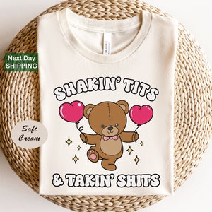 May include: Soft cream-colored t-shirt with the text "SHAKIN' TITS & TAKIN' SHITS" in a playful font. A cartoon teddy bear holds two pink heart-shaped balloons. The shirt also has the text "Next Day SHIPPING".