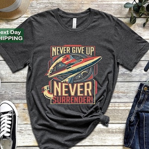 May include: Dark gray t-shirt featuring a retro spaceship graphic with the words "NEVER GIVE UP NEVER SURRENDER!" in a vintage style. The design includes red, blue, and yellow accents, evoking a classic sci-fi aesthetic.