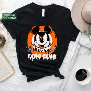 May include: Black t-shirt featuring a Halloween-themed Mickey Mouse design. The graphic includes Mickey's face within an orange circle, with the text "Mickey Mouse Fang Club" in a spooky font. The shirt is styled on a white surface.