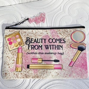 May include: A rectangular makeup bag with a black zipper and a pink glitter charm. The bag features illustrations of makeup items like lipstick, eyeshadow, and a brush, with the text "BEAUTY COMES FROM WITHIN (within this makeup bag)".