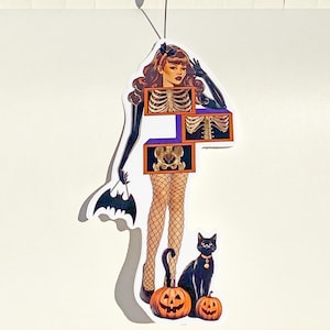 Puede incluir: Una pegatina con temática de Halloween que presenta a una mujer con torso de esqueleto, guantes negros y medias de rejilla. Un bolso con forma de murciélago, dos calabazas talladas y un gato negro con collar completan el diseño. La pegatina está adherida a una superficie blanca.