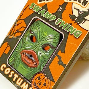 May include: A Halloween costume box featuring a green Swamp Thing mask with orange eyes and a red mouth. The box is orange and black with the word "Halloween" and "Costume" printed on it. A jack-o'-lantern is also on the box.