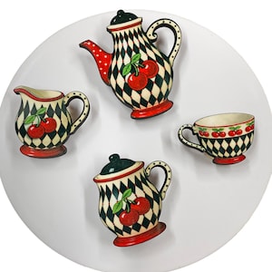 Puede incluir: Un conjunto de cuatro elementos decorativos de cocina de madera: una tetera, una jarra de crema, un azucarero y una taza. Cada pieza presenta un patrón de diamantes en blanco y negro, detalles en rojo y un motivo de cerezas. Dispuestos sobre una superficie circular blanca.