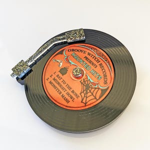 May include: A black vinyl record with a glittery silver and black tone arm. The record label is orange with the text "Groove Witch Records Presents Haunted Hits". Song titles include "Bat to the Bone" and "Monster Mash".