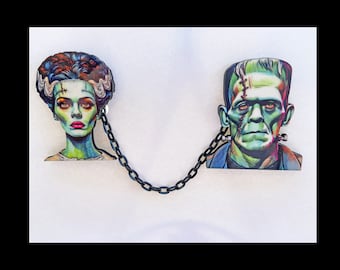 May include: A pair of black and white illustrated portraits of Frankenstein's monster and his bride connected by a black chain. The portraits are on a white background.