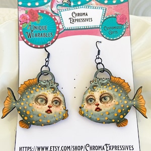 May include: A pair of earrings featuring a whimsical, cartoon-style fish with yellow and blue polka dots. The fish has a large, round head with big eyes and a pink mouth. The earrings are made of wood and have black metal hooks. The earrings are displayed on a white card with the text "Chroma Expressives" and a website address.
