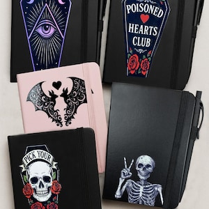 Puede incluir: Una colección de cuadernos negros con diseños únicos. Uno presenta un ataúd con un ojo y lunas crecientes, otro dice "Poisoned Hearts Club", y un tercero tiene una calavera con rosas. También se incluye un cuaderno rosa con un diseño de murciélago.