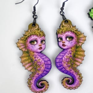 May include: A pair of whimsical seahorse earrings. Each earring features a purple seahorse with gold and pink accents, detailed with large eyes and a small red mouth. The earrings hang from black metal hooks.