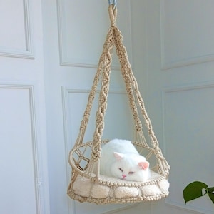 Puede incluir: Un gato blanco descansando en una hamaca de macramé beige para gatos colgada del techo.
