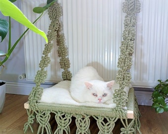 Può includere: Un gatto bianco che riposa in un'amaca per gatti in macramè con una frangia verde. L'amaca è appesa a un termosifone bianco.