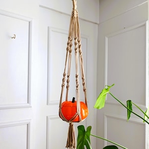 Pode incluir: Um suporte de plantas de macramé com um vaso em forma de abóbora pendurado nele. O suporte é feito de corda de juta natural e tem uma borla na parte inferior. O vaso é laranja e tem um acabamento liso e brilhante.