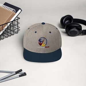 May include: A gray and navy blue baseball cap with the words "Interpreting Sign World" embroidered on the front. The cap features a circular logo with red, yellow, and blue elements. A pair of headphones and a notebook are in the background.