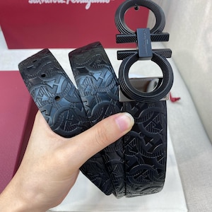 May include: Black leather belt with a textured pattern and a large, black buckle. The buckle features a double Gancini design. The belt is displayed against a red box with the Salvatore Ferragamo logo.