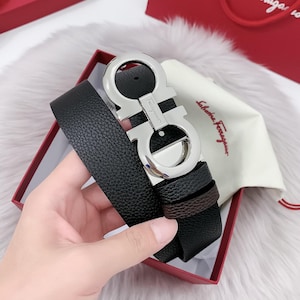 Puede incluir: Cinturón de cuero negro con hebilla plateada. La hebilla tiene un diseño en forma de doble G. El cinturón se muestra en una caja roja, con una tela blanca y una bolsa de compras roja en el fondo.