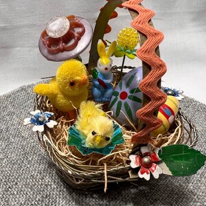 May include: A vintage metal Easter basket filled with a variety of decorations, including two yellow chicks, a blue bunny, a purple flower, and three decorated eggs. The basket has a pink scalloped handle.