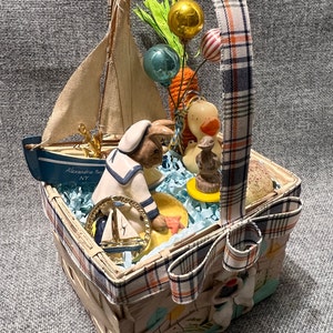 May include: A wicker basket with a plaid bow and a blue and white sailor bunny figurine inside. The basket is filled with blue and white tissue paper and other Easter decorations, including a small sailboat, a carrot, and a yellow duck.