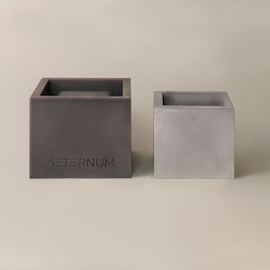 May include: Two square concrete planters, one grey and one dark grey. The dark grey planter has the word "AETERNUM" embossed on the front.
