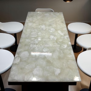 Op de afbeelding: Een rechthoekige eettafel met een lichtgroen en wit patroon, omringd door zes witte stoelen met zwarte poten. Het tafelblad heeft een glanzende afwerking. De stoelen hebben een modern design.