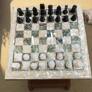 May include: A chess set featuring a square board inlaid with mother-of-pearl and black and white chess pieces. The board is supported by a small wooden stand. The chess pieces are in their starting positions.