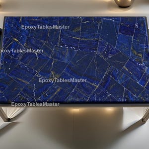 May include: Rectangular coffee table with a dark blue, mosaic-style top. The surface is made of irregularly shaped pieces with gold veining. The table has a dark metal frame and is surrounded by blue chairs. The text "Epoxy TablesMaster" is visible on the table.