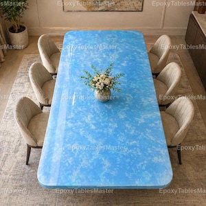 Pode incluir: Uma grande mesa de jantar retangular com uma superfície brilhante de resina epóxi azul claro. A mesa é rodeada por seis cadeiras estofadas bege. Um pequeno vaso de flores brancas está no centro. A mesa mede cerca de 254 cm de comprimento.