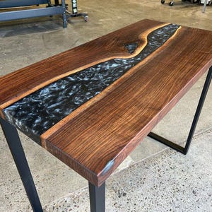 May include: A rectangular wooden table with a dark walnut finish and a black epoxy resin river running through the center. The table has black metal legs and is suitable for a living room or office.