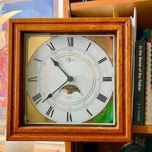 May include: Square wooden-framed clock with a white face, black Roman numerals, and gold accents. The clock face has a moon phase indicator and the word "Elgin". The clock is surrounded by books and artwork.