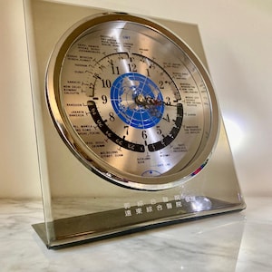 May include: A square-shaped world time clock with a silver metallic finish and a circular face. The dial features a blue map of the world, black hour numerals, and a rotating outer ring listing global cities. Chinese text is printed at the bottom.