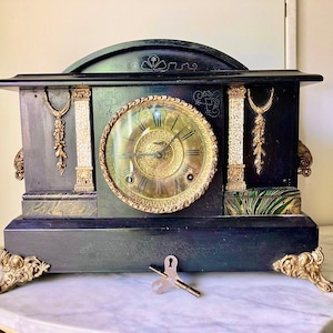 Puede incluir: Un reloj de sobremesa negro con una esfera circular dorada que presenta números romanos. La caja del reloj está decorada con detalles de metal dorado, incluyendo pies ornamentados, columnas laterales y motivos florales. Una llave de cuerda está frente al reloj.