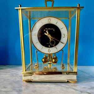 May include: A rectangular, gold-framed clock with a glass enclosure. The clock face is white with black center and gold hands. The pendulum has two gold-colored spheres. The clock sits on a white base.